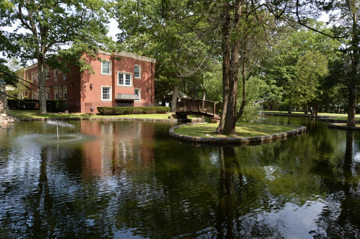 A serene scene featuring a brick building by a pond, surrounded by lush trees and a small bridge.