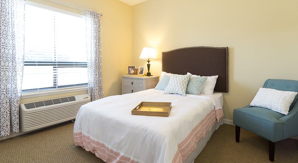 A cozy bedroom with a neatly made bed, soft lighting from a table lamp, and a nightstand by the window.