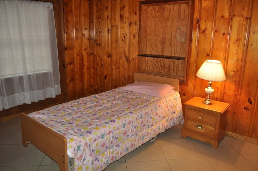 A cozy bedroom with wood-paneled walls, featuring a single bed covered with a patterned quilt, next to a nightstand with a lamp.