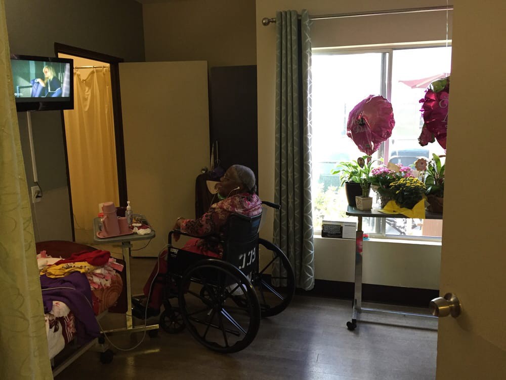 A person in a wheelchair watching TV in a hospital room adorned with flowers by the window.