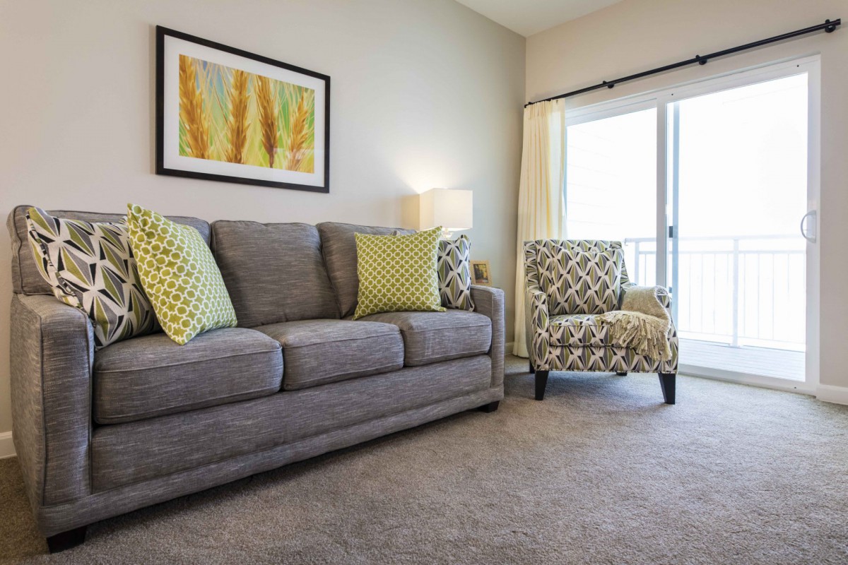 A cozy living room with a gray sofa and an armchair, decorated with yellow and green pillows, featuring artwork on the wall.