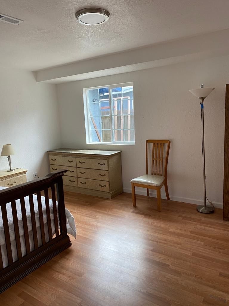 A tidy bedroom with a wooden floor, featuring a dresser, chair, bed, and standing lamp near a window.