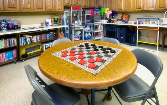 A game room featuring a round table set up for a checkers match with shelves of toys and games in the background.