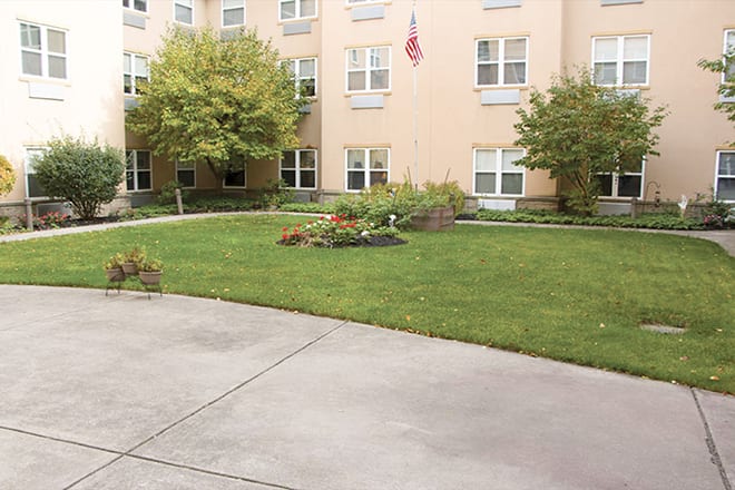 A well-maintained garden in front of a multi-story building with a lawn, trees, and an American flag.