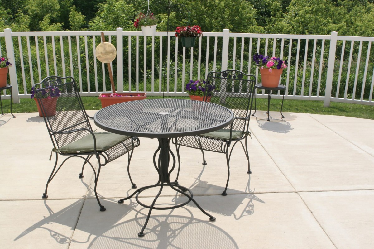 A sunny patio with a glass-top table and metal chairs, surrounded by potted plants and flowers.