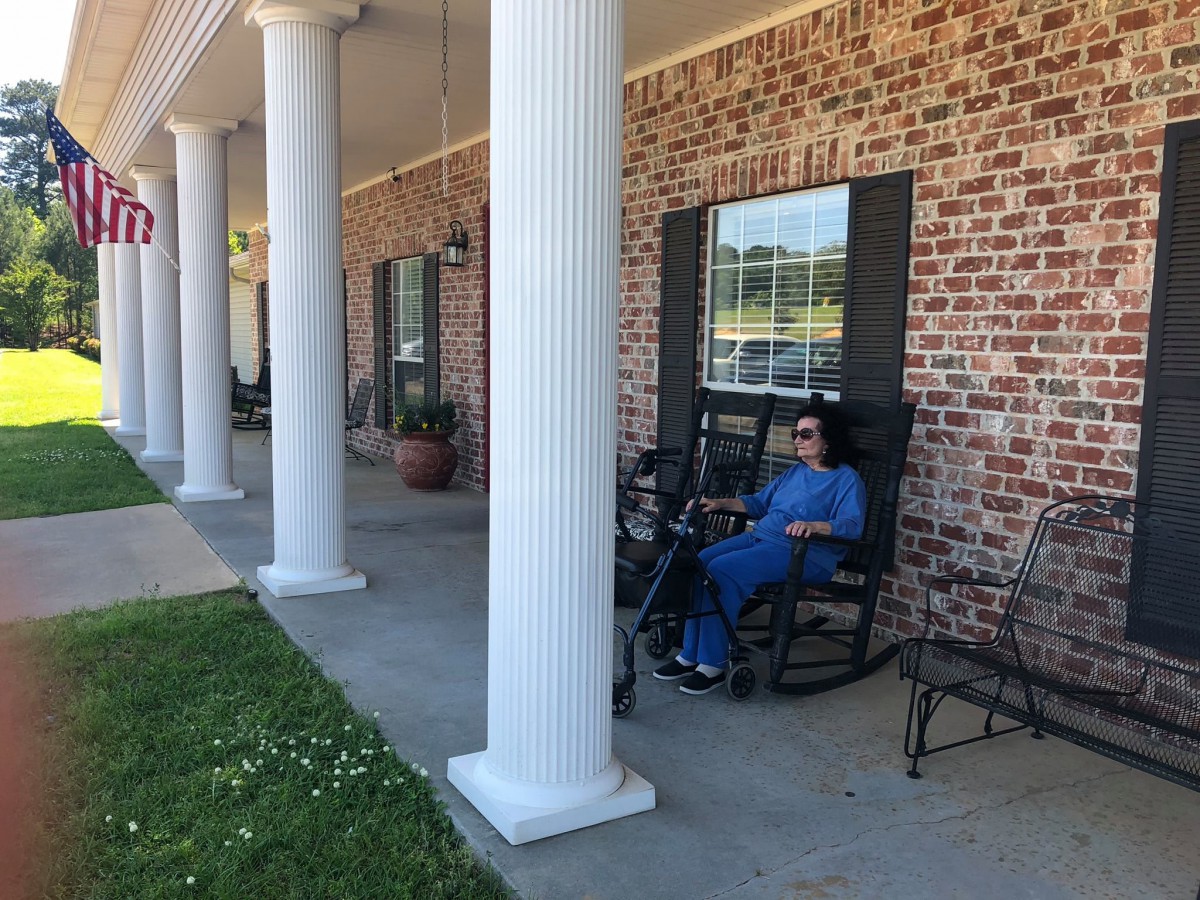 A person in blue attire relaxes on a rocking chair on a porch with large white columns and a brick facade.