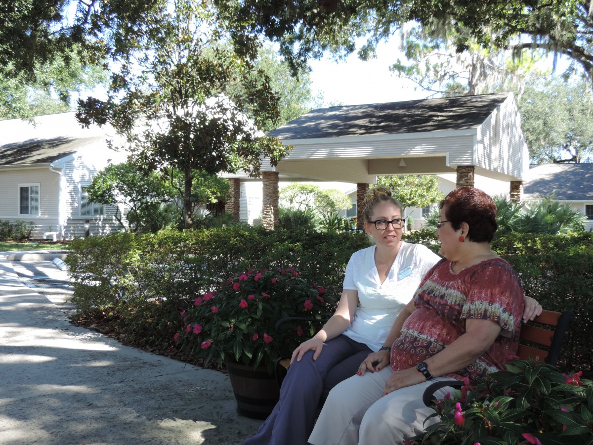 Two people sitting on a bench in a lush garden enjoying a conversation under the shade of trees.