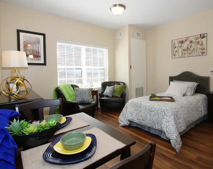 A cozy bedroom with a single bed and an adjoining living space, featuring armchairs and a small dining area.