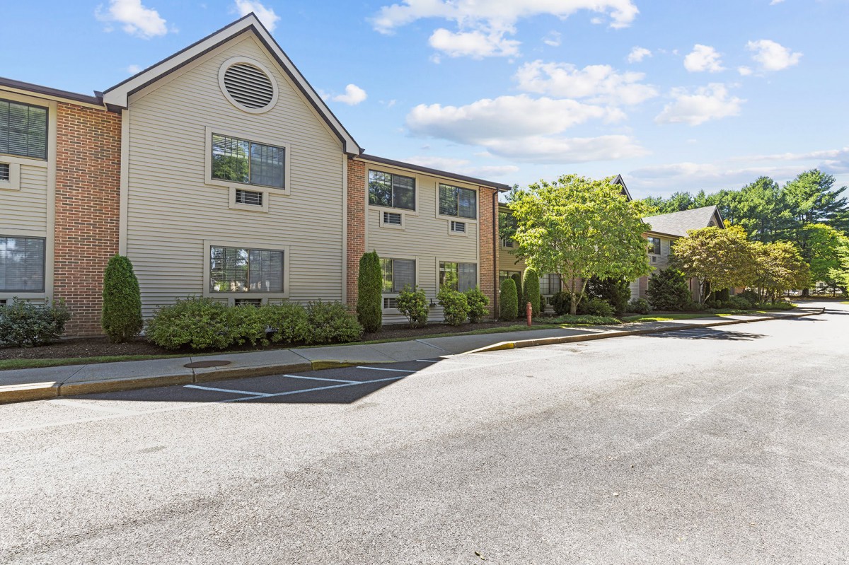 A view of a residential building with a parking lot and trees on a sunny day.