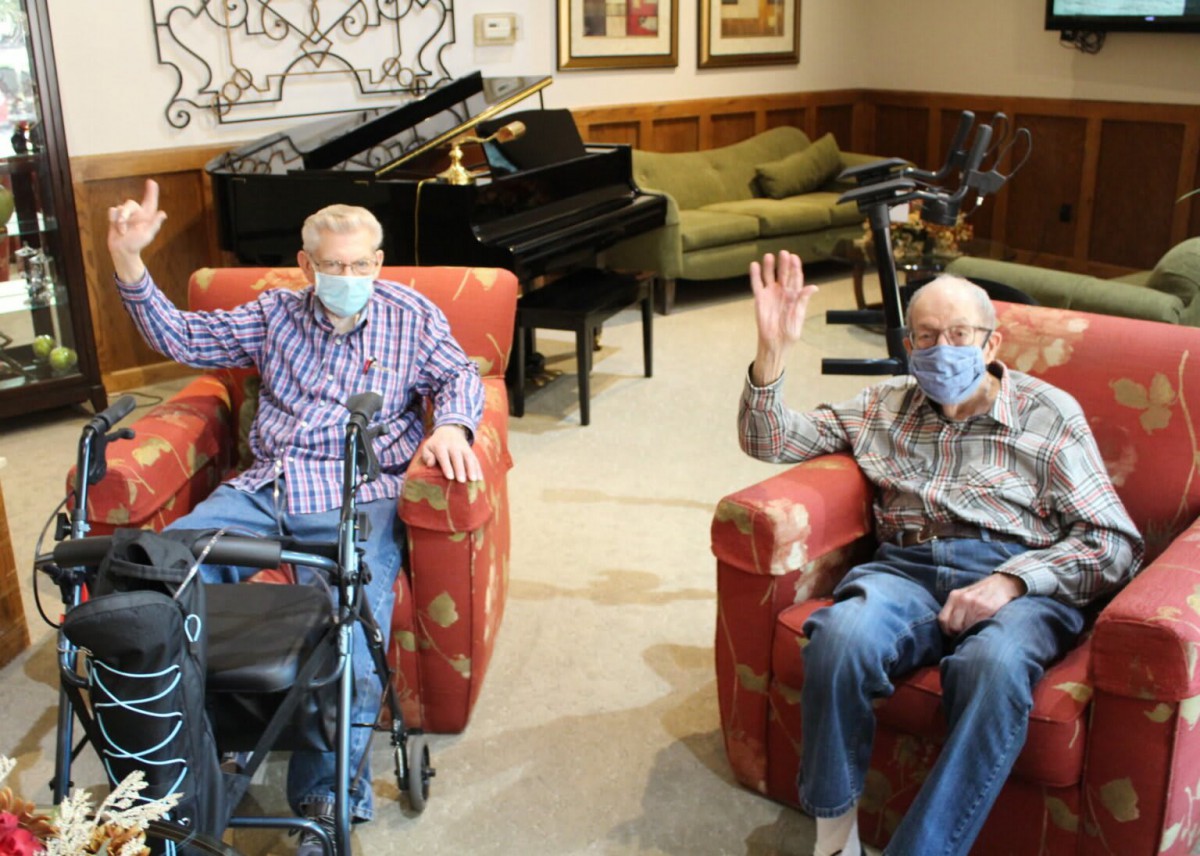Two elderly individuals wearing masks, sitting in a living room setting with a piano in the background.