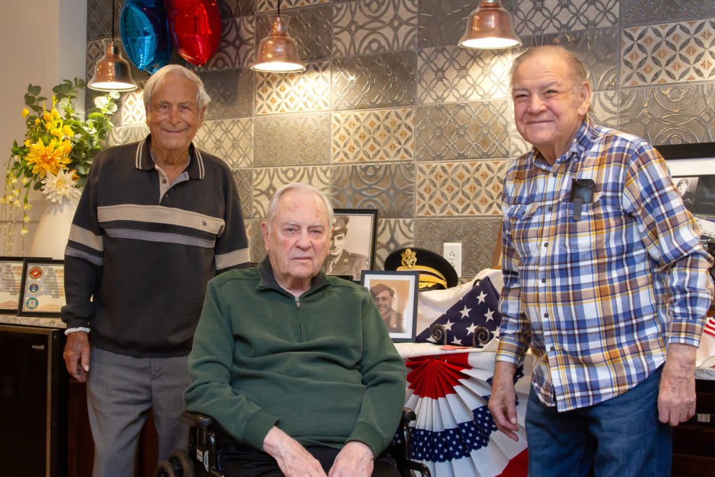 Three elderly men gathered indoors, surrounded by balloons and a flag for a celebration.