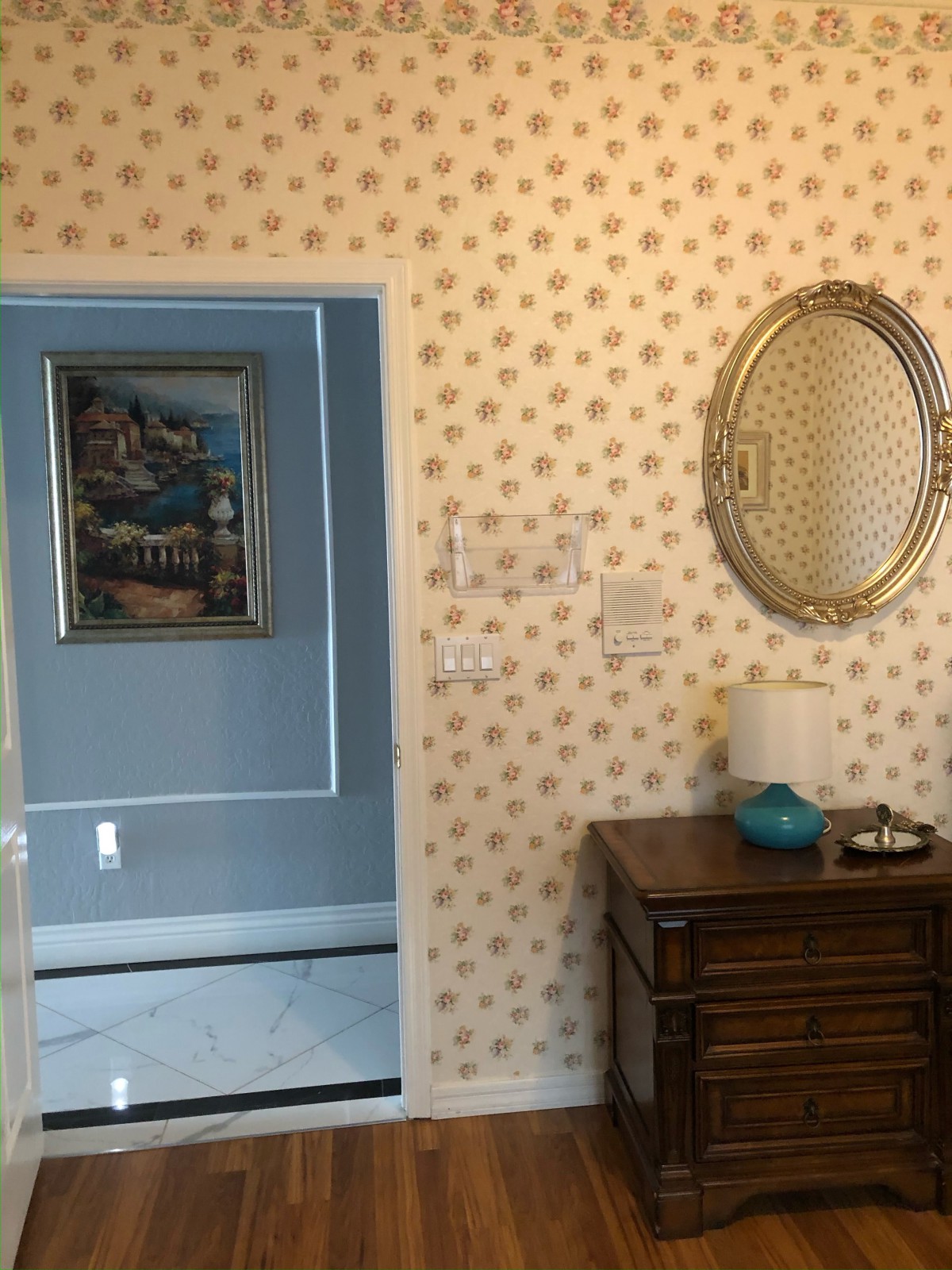 A cozy interior featuring floral wallpaper, a wooden drawer with a lamp and decorative items, an oval mirror on the wall, and a framed painting in the adjacent room.