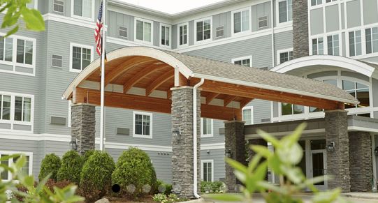 A modern building entrance with stone pillars and a wooden arch, featuring a flag and landscaped greenery.