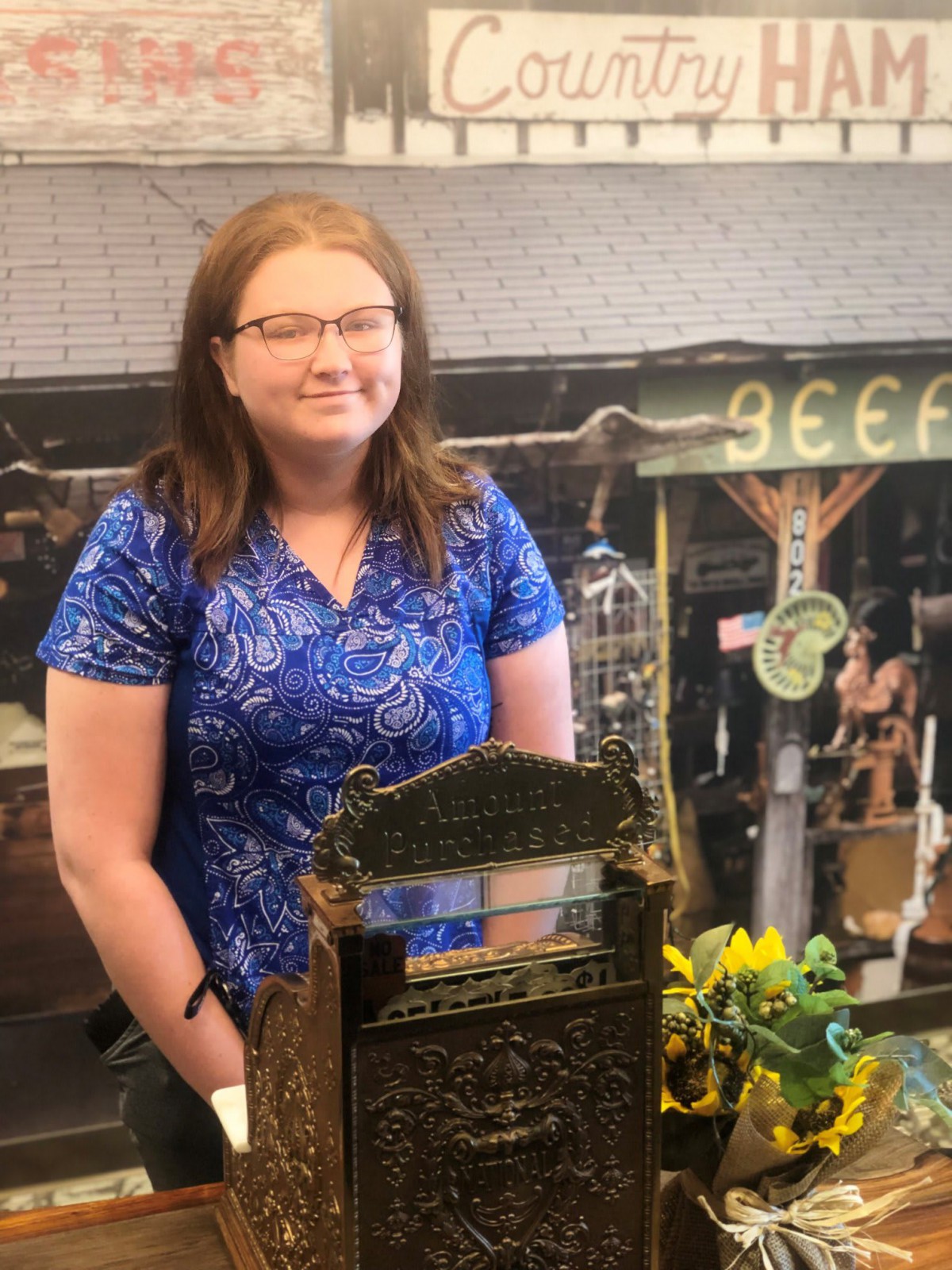 A woman with glasses stands next to an antique cash register indoors.
