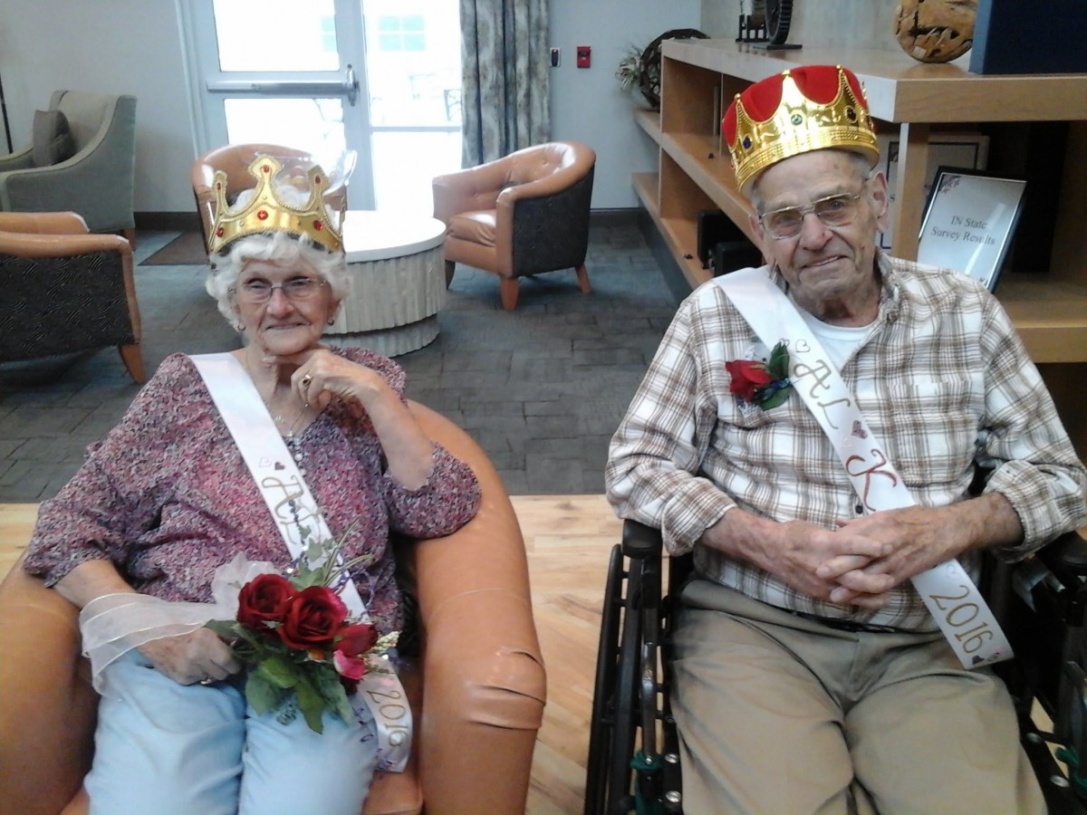 An elderly man and woman wearing crowns and sashes, holding flowers, sitting together indoors.