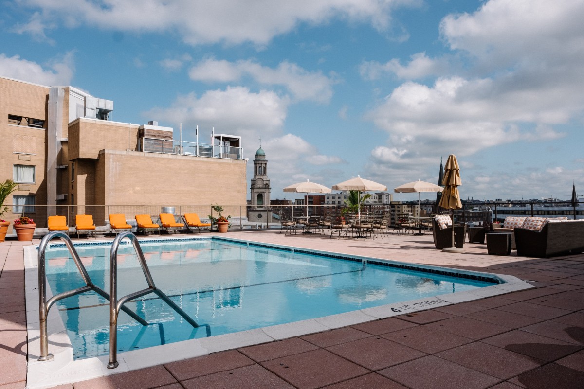 A rooftop swimming pool with deck chairs overlooking the city skyline under a partly cloudy sky.