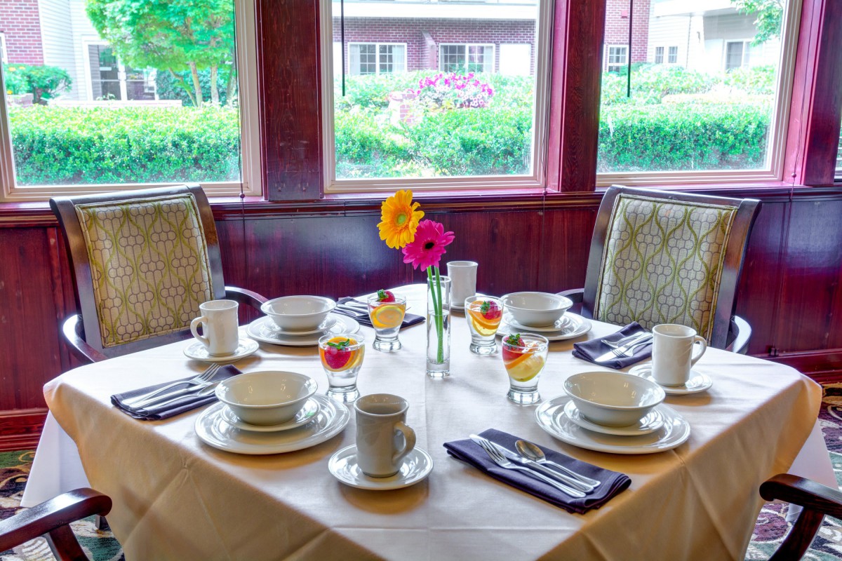 A beautifully set dining table with cups, bowls, and floral centerpieces by a large window.