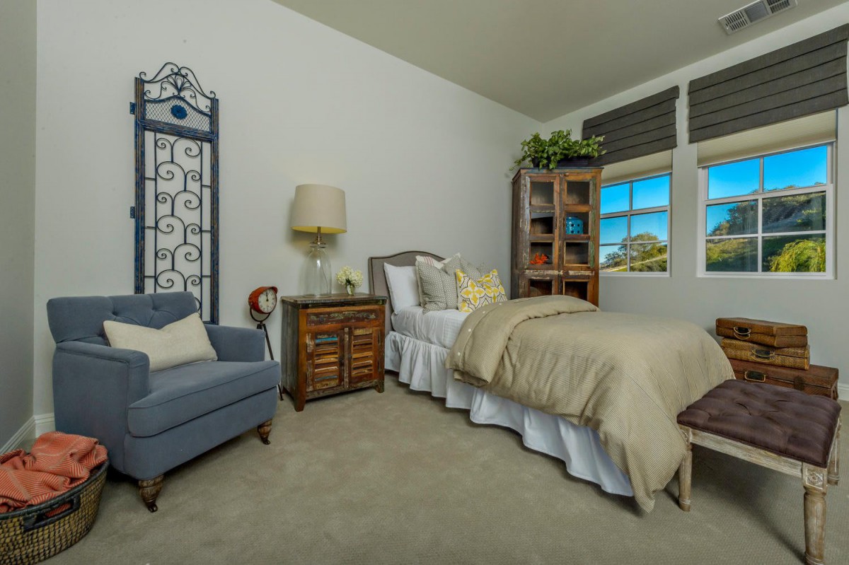 A cozy bedroom featuring a bed with neutral bedding, a blue armchair, and a wooden nightstand under a lamp.