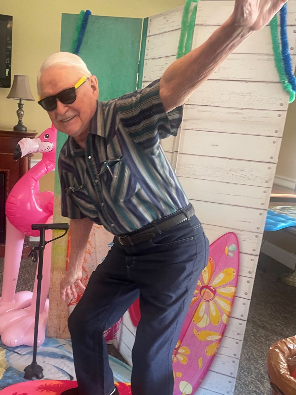 An elderly man posing playfully with a flamingo prop and colorful decorations.
