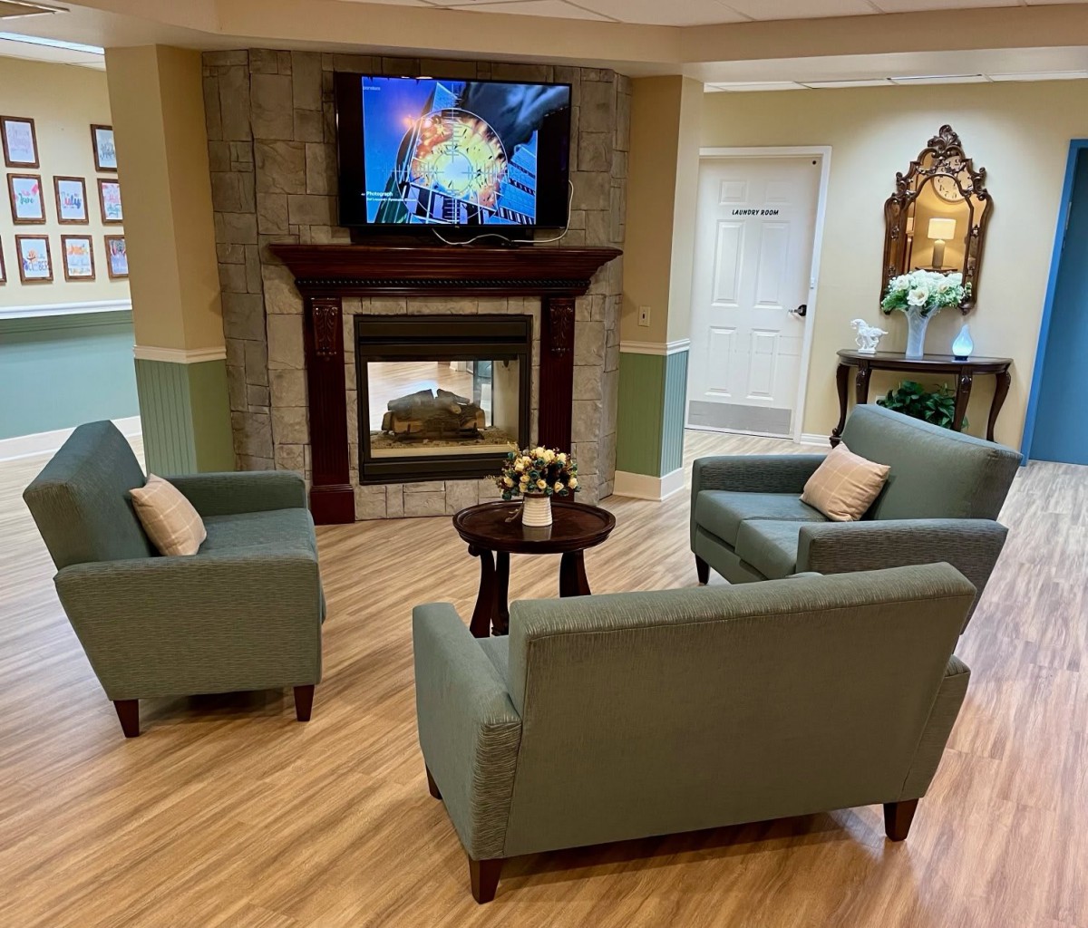 A cozy living room with green sofas arranged around a central table, featuring a fireplace and a wall-mounted television.