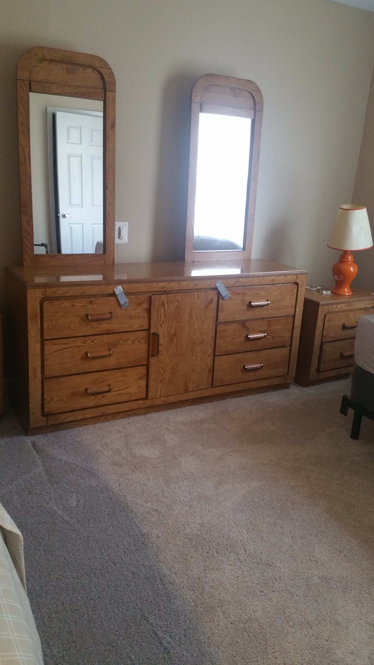 A wooden dresser with attached mirrors and a lamp on the side table in a bedroom.