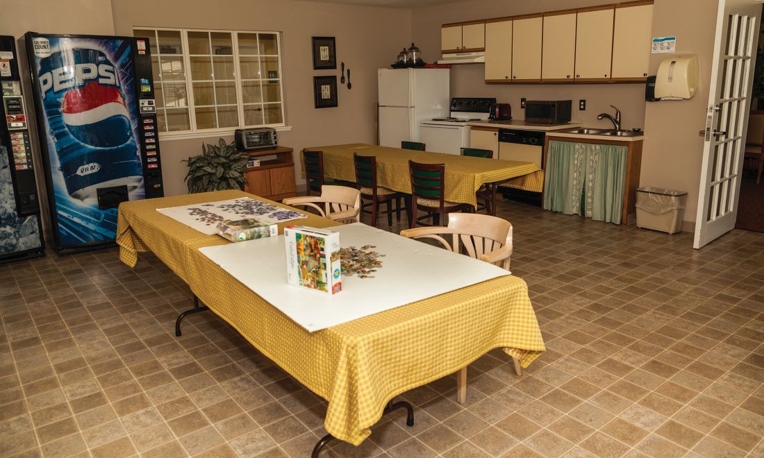 A spacious kitchen and dining area with tables covered in yellow tablecloths, chairs, a refrigerator, and kitchen cabinets.
