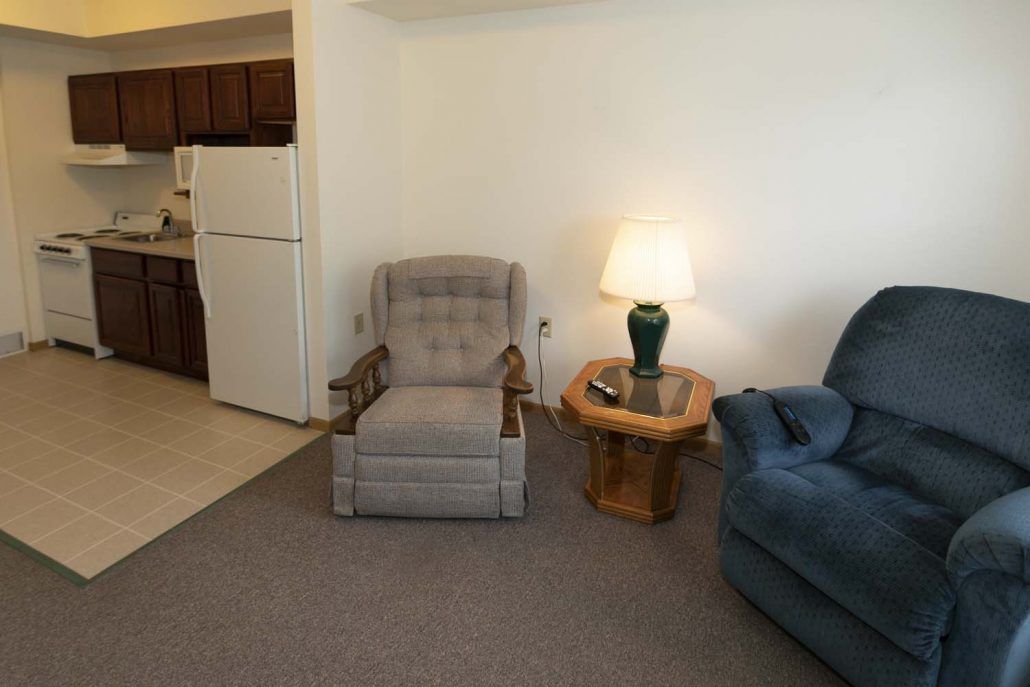 A cozy living room with a gray armchair, a wooden side table with a lamp, and a blue sofa. The kitchen area is visible in the background.