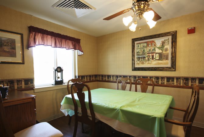 A cozy dining room with a table covered by a green cloth, surrounded by wooden chairs. A painting hangs on the wall, and a ceiling fan with lights is overhead.