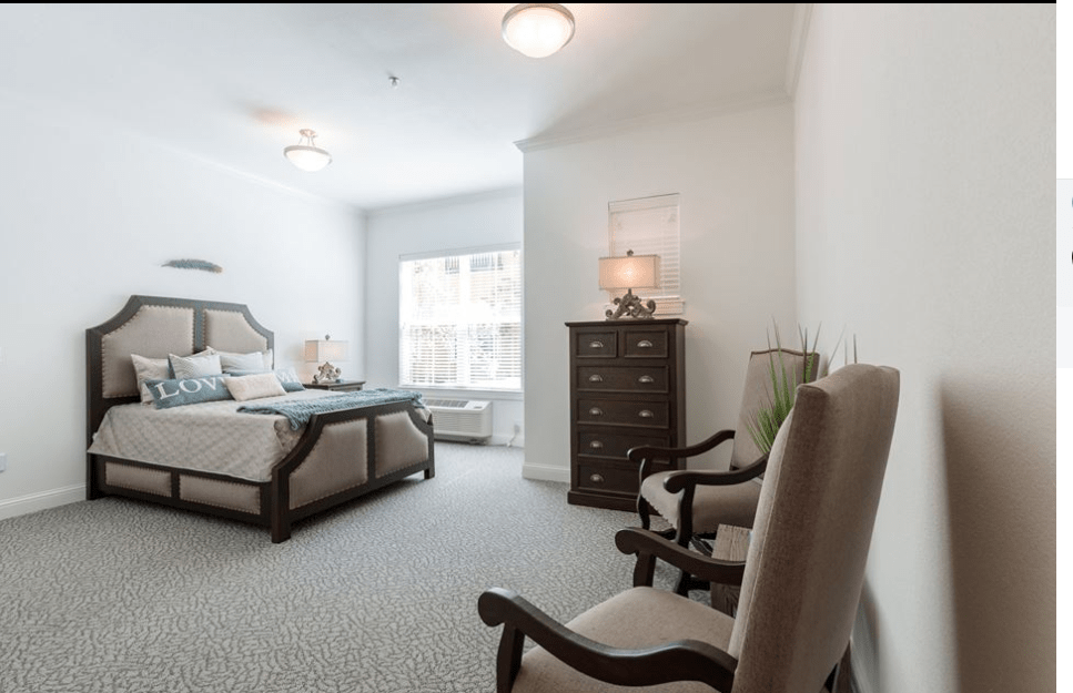 A spacious and elegantly furnished bedroom featuring a bed, dresser, chairs, and a large window allowing natural light.