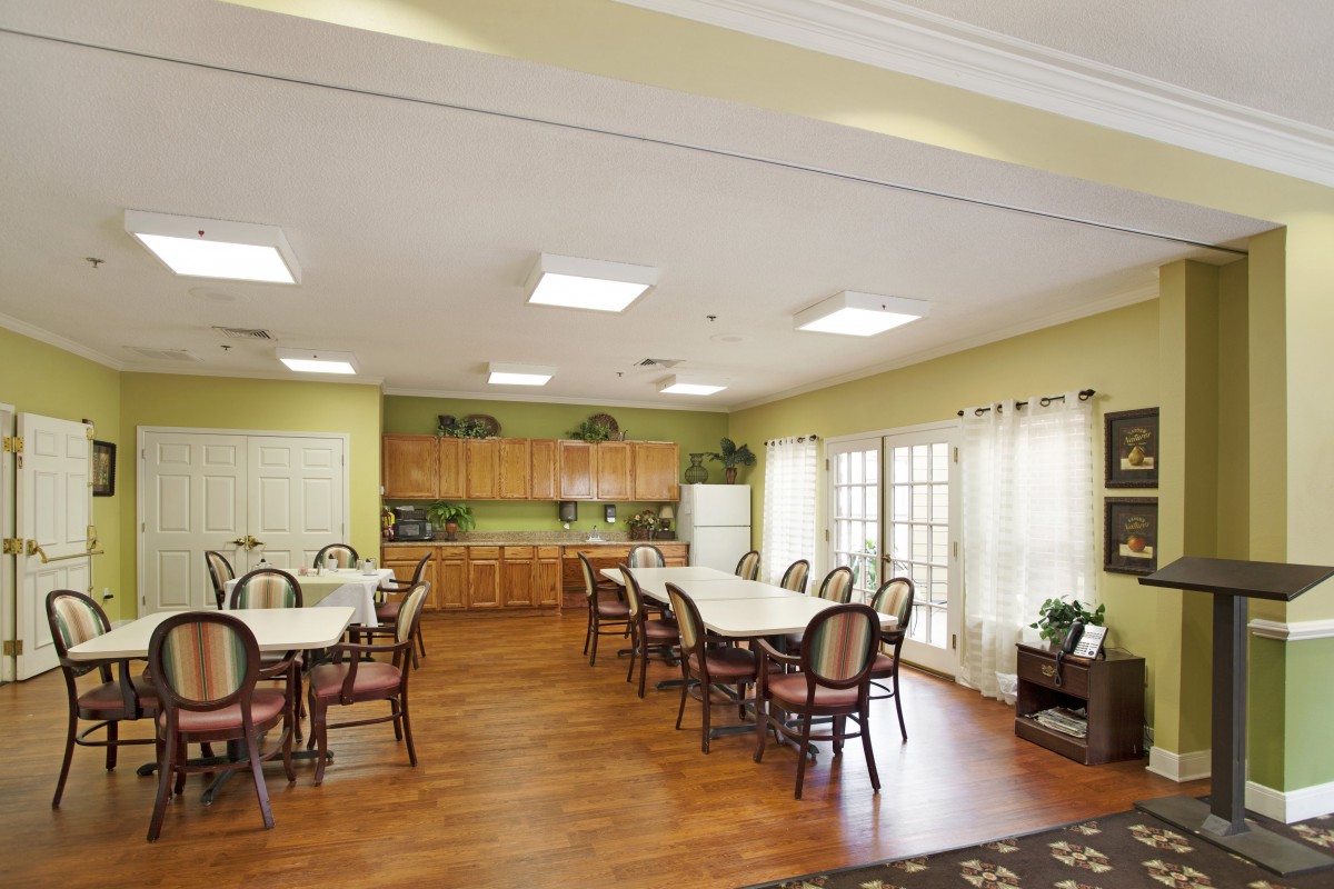 A spacious dining room with multiple tables and chairs, adjacent to a small kitchen area, featuring hardwood flooring and large windows providing plenty of natural light.