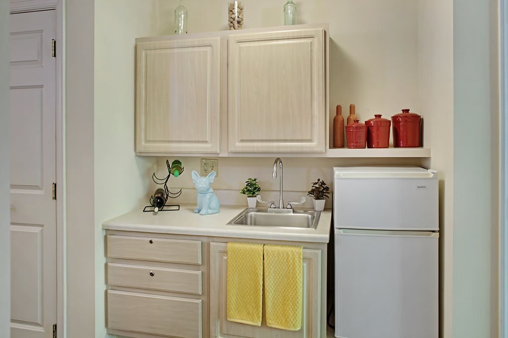 A small kitchen area featuring cabinets, a sink with yellow towels, a refrigerator, and decorative items on the countertop.