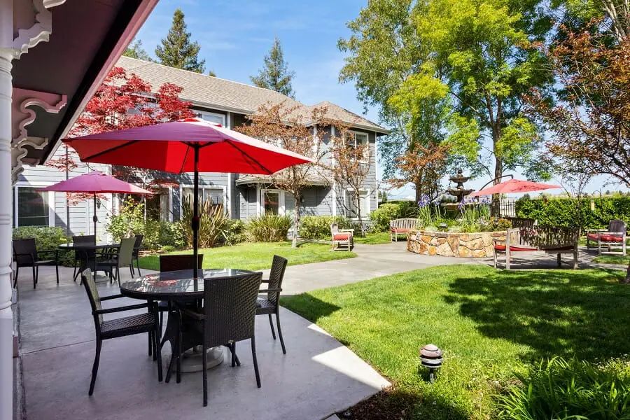 A sunny outdoor patio area with tables, chairs, and red umbrellas surrounded by a lush garden and trees.
