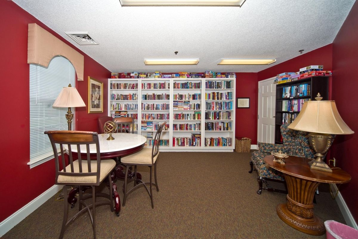 A cozy library room with red walls, featuring bookshelves filled with books, a round table and chairs, and several lamps for lighting.