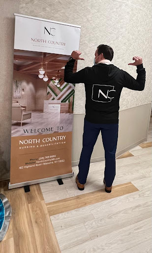 A person showcasing a hoodie with a logo, standing next to a welcoming banner for an event.