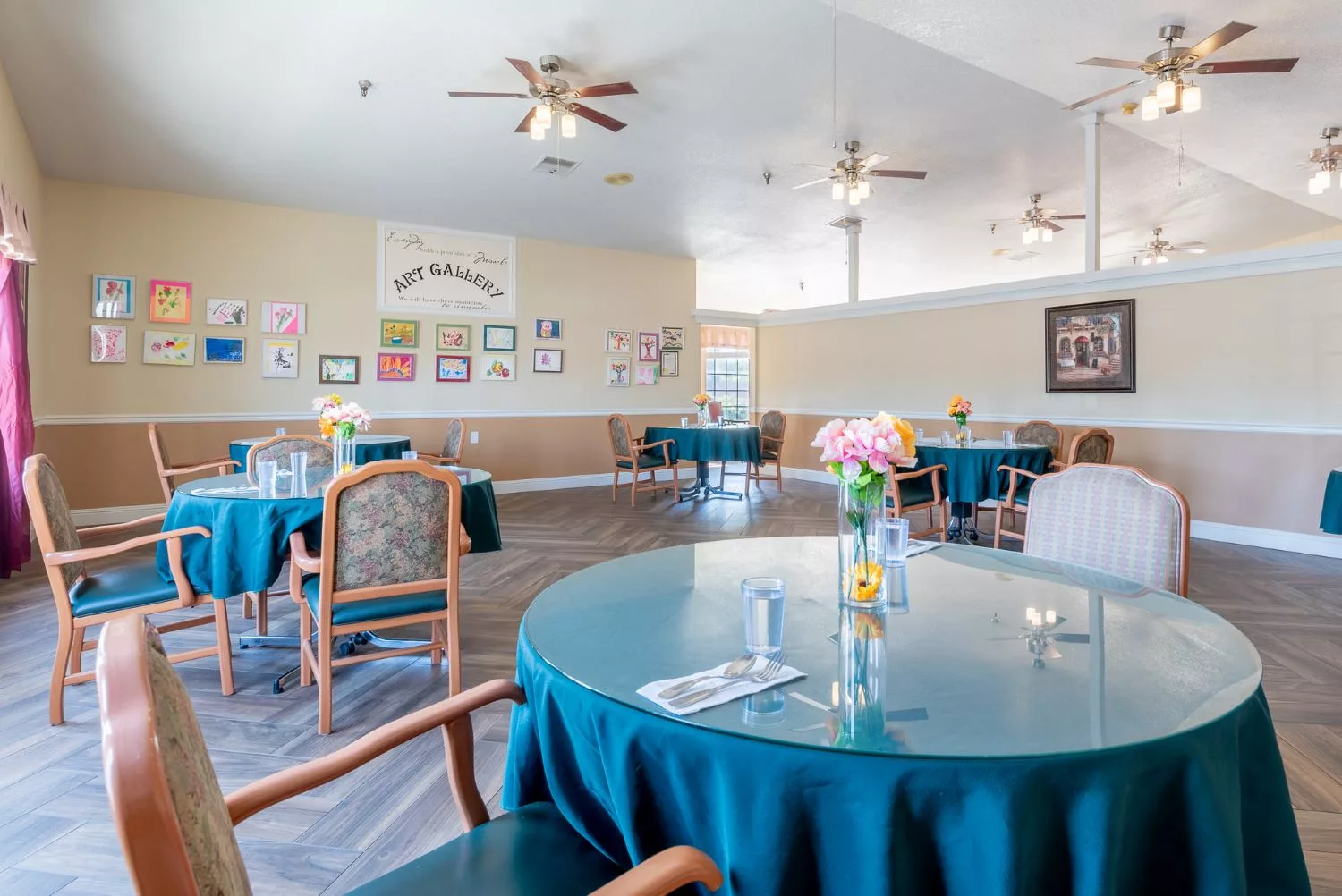 Spacious dining room with round tables, decorated with flowers and ceiling fans overhead.
