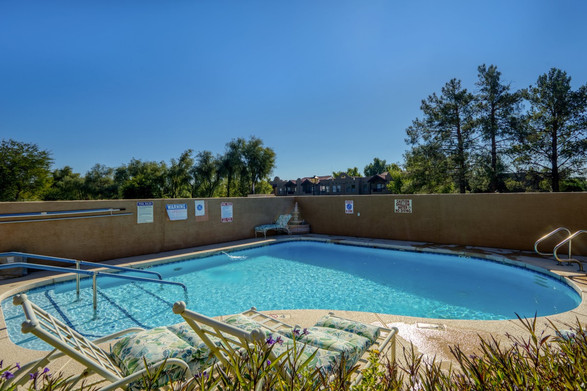 An outdoor swimming pool surrounded by a fence with trees in the background under a clear blue sky.
