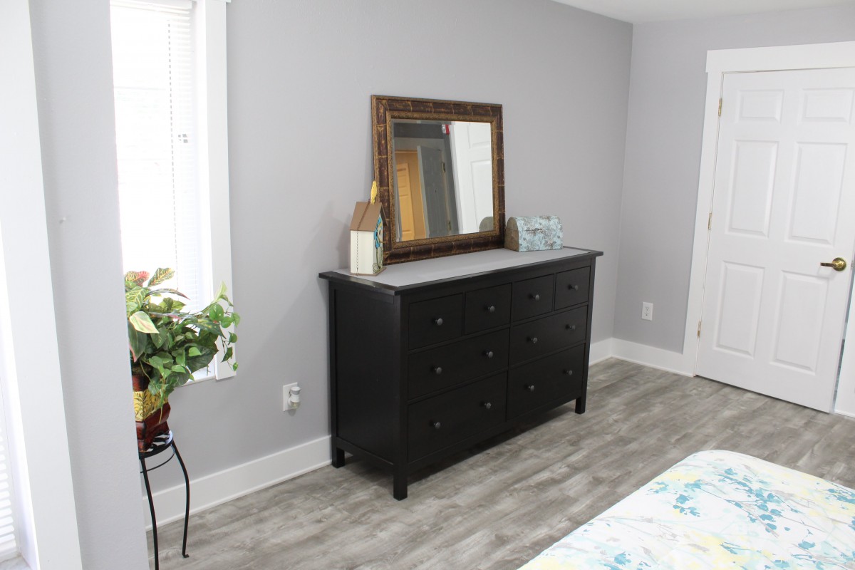 A bedroom interior featuring a dark dresser with a mirror above, decorated with plants and accessories.