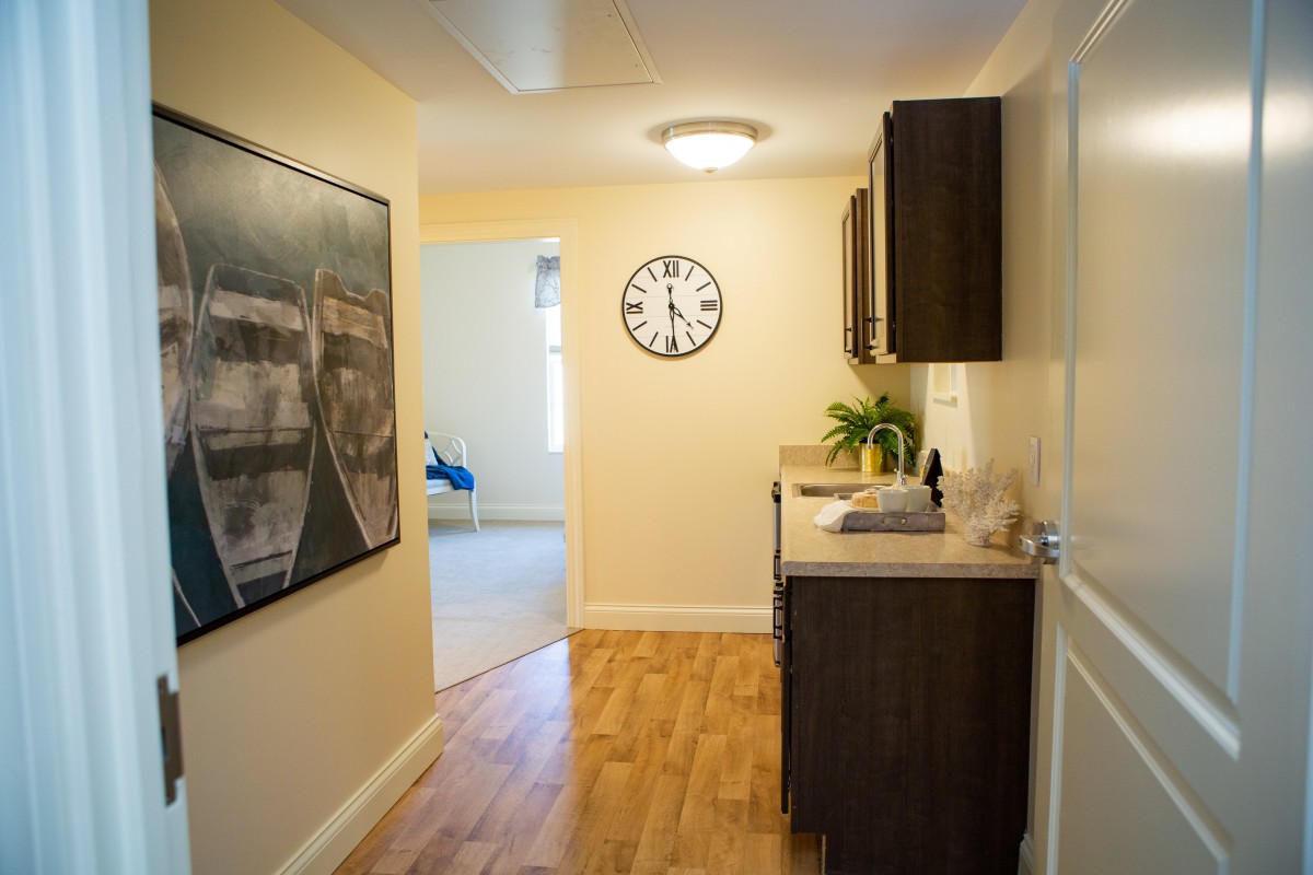 A well-lit hallway with wooden flooring, featuring a wall-mounted clock and artwork, along with dark cabinets for storage.