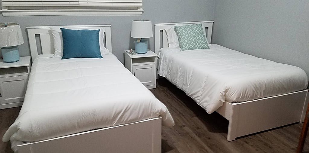 A cozy bedroom with twin beds, each adorned with white bedding and colorful pillows, separated by a nightstand with a lamp.