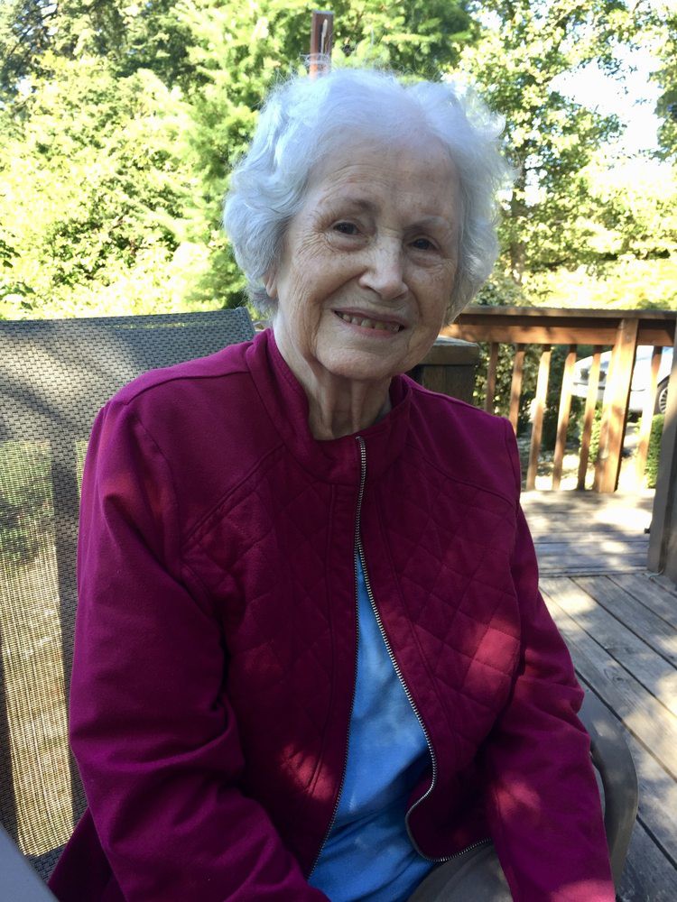 An elderly woman smiles while sitting outdoors in a red jacket.