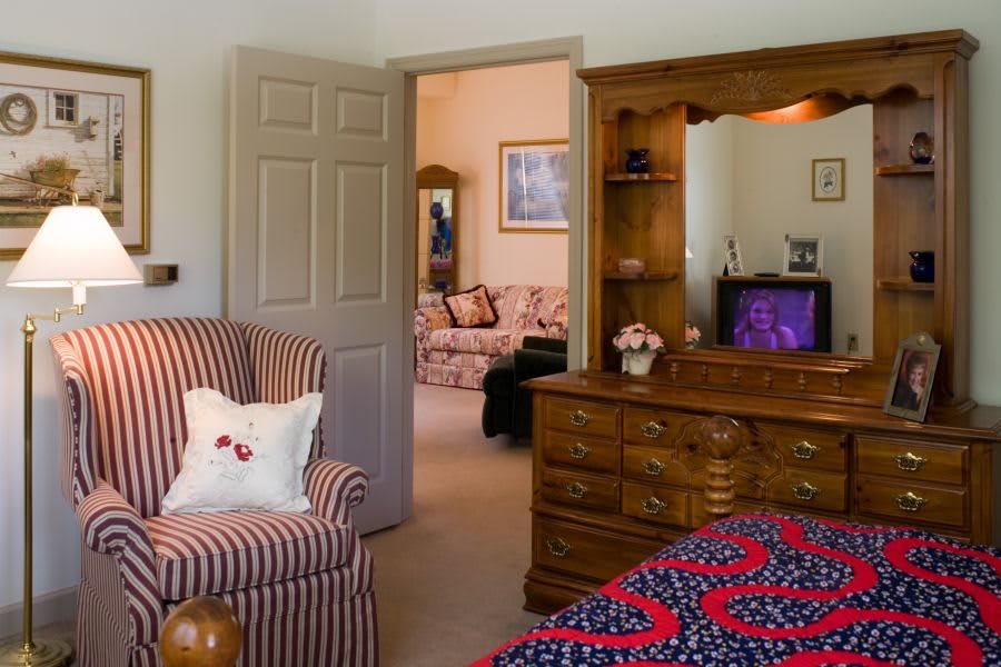 A cozy bedroom featuring a dresser with a mirror and television, a striped armchair with a decorative pillow, and a colorful quilt on the bed.