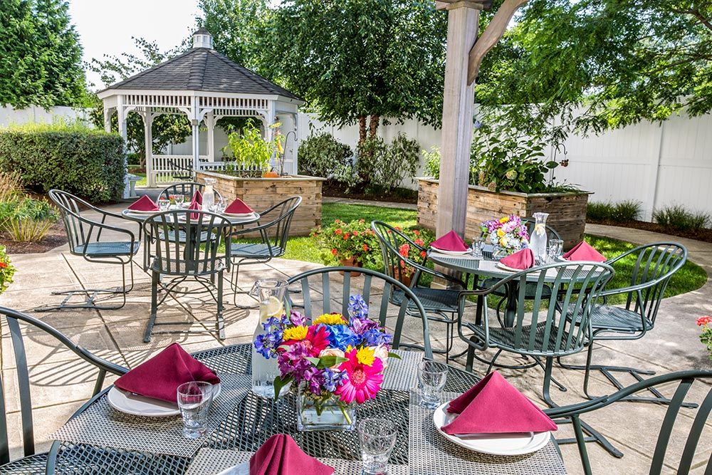 A beautifully set outdoor dining area with tables adorned by vibrant flowers and burgundy napkins, surrounded by a lush garden and a charming gazebo.