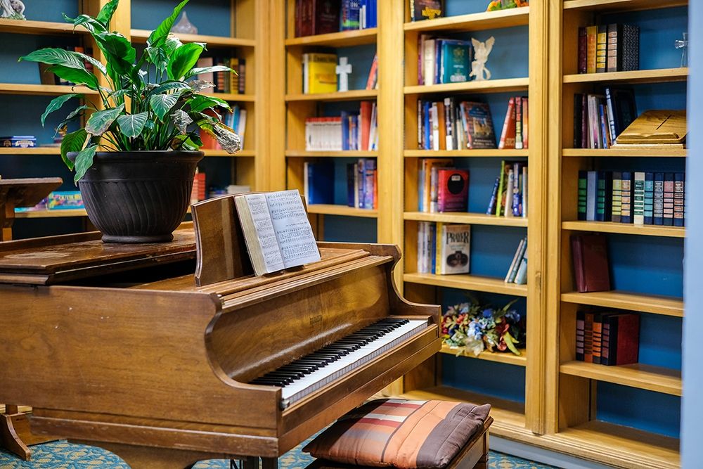 A cozy library corner featuring a piano with sheet music, surrounded by bookshelves and a potted plant.