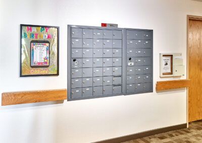 A set of gray mailboxes mounted on a wall in a hallway, accompanied by bulletin boards on either side.