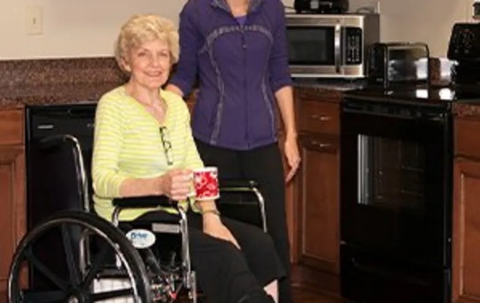 An elderly woman in a wheelchair holding a mug, with a caregiver standing beside her in the kitchen.
