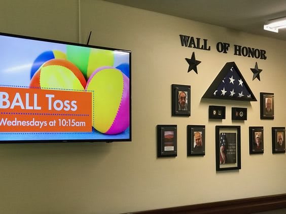 A 'Wall of Honor' display featuring framed photos and a folded flag, with a television advertising a ball toss event.