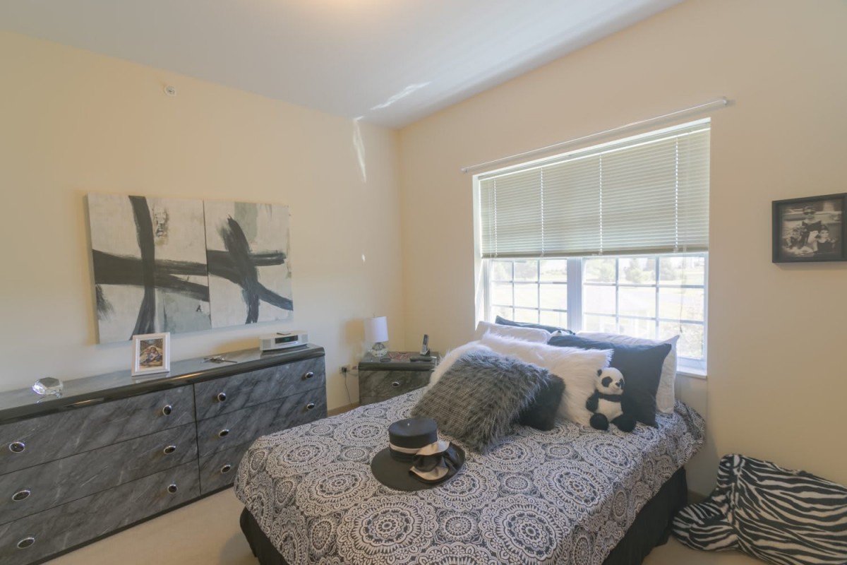 A cozy bedroom with a neatly made bed, decorative pillows, a dresser, and a large window letting in natural light.