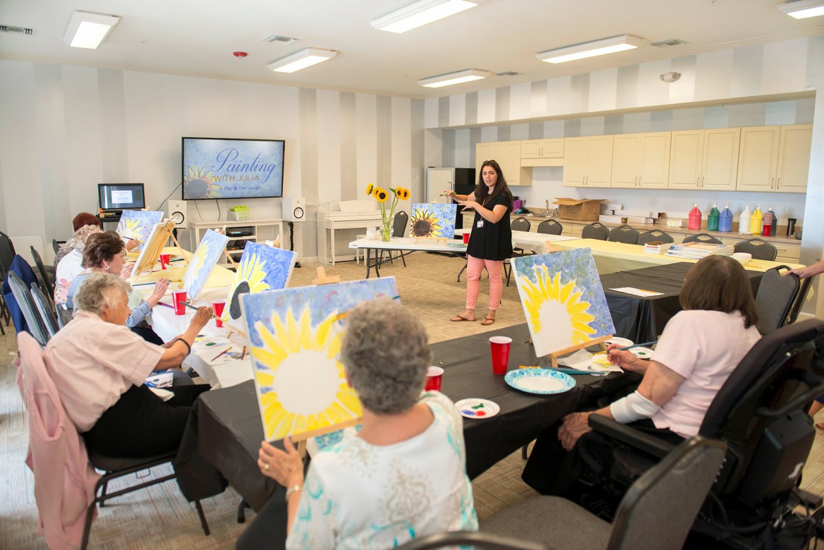 A group of adults participating in a painting workshop, each working on their own canvas while an instructor guides the session.