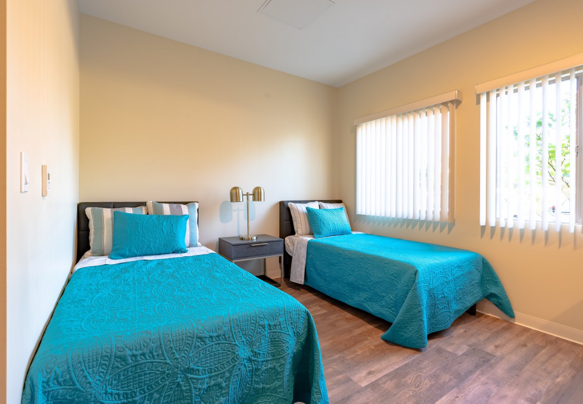 A cozy bedroom with twin beds featuring turquoise bedding, illuminated by natural light from the windows.