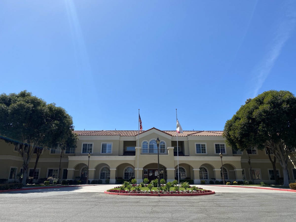 A large building with arched windows and flags under a clear blue sky, surrounded by trees and landscaped greenery.